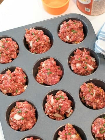 A muffin tin filled with raw meatloaf mixture ready to be baked. The meatloaf muffin recipe portions appear roughly shaped and are topped with bits of onion and herbs. An orange bowl and a tin can are visible in the background.