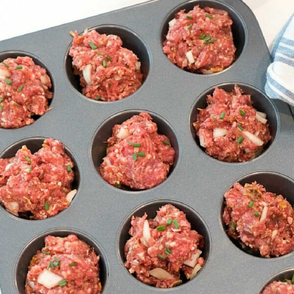 A muffin tin filled with raw meatloaf mixture ready to be baked. The meatloaf muffin recipe portions appear roughly shaped and are topped with bits of onion and herbs. An orange bowl and a tin can are visible in the background.