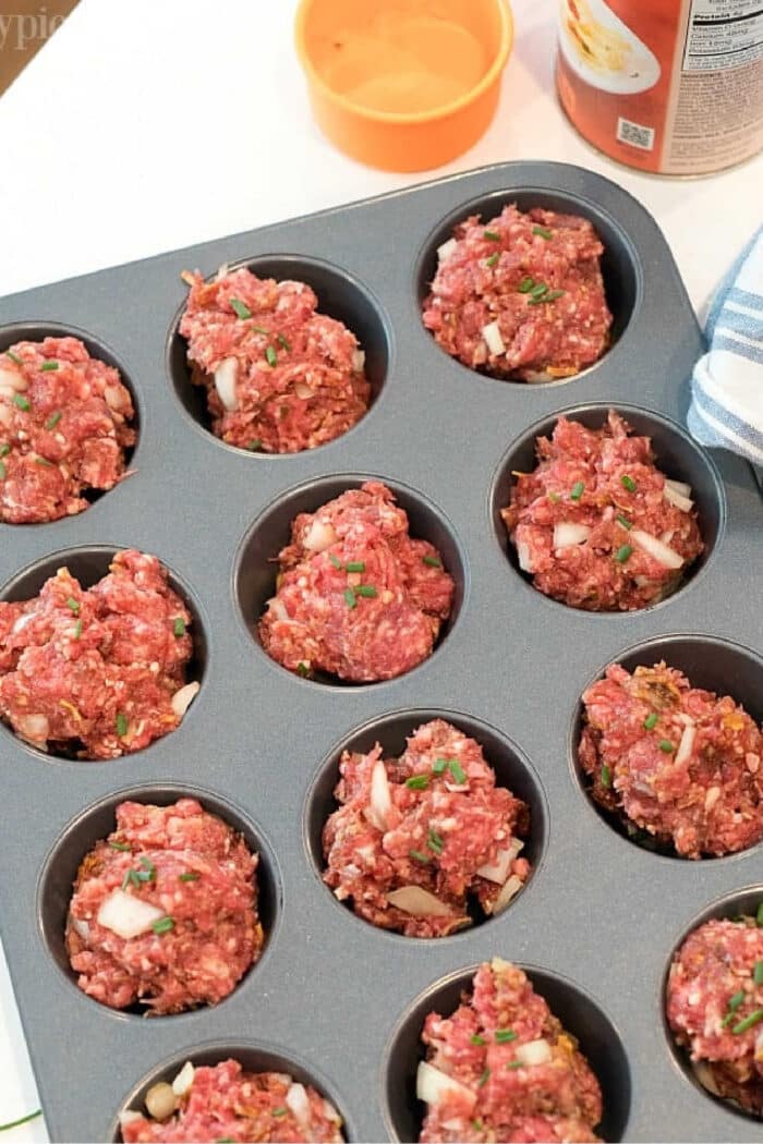 A muffin tin filled with raw meatloaf mixture ready to be baked. The meatloaf muffin recipe portions appear roughly shaped and are topped with bits of onion and herbs. An orange bowl and a tin can are visible in the background.