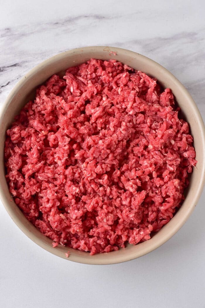 A bowl filled with raw ground meat rests on a marble surface, inviting you to ponder how long to cook ground beef for the perfect savory dish.