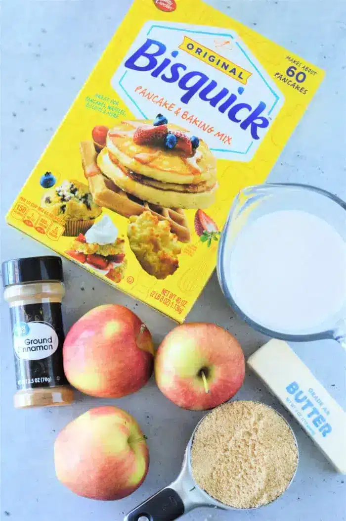 A box of Bisquick Pancake and Baking Mix is surrounded by a measuring cup with milk, a bottle of ground cinnamon, three apples, a stick of butter, and a measuring cup filled with brown sugar on a light-colored surface—everything you need for an easy Apple Cobbler with Bisquick.