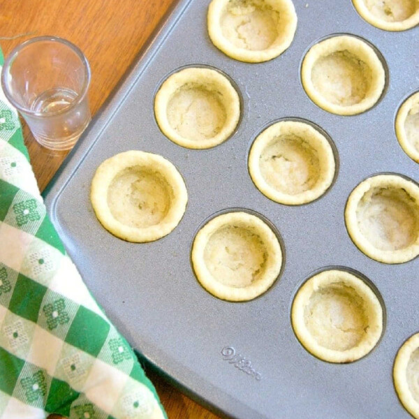 Mini cookie cups in a mini muffin pan beside a green checkered oven mitt and a small glass on the wooden surface.