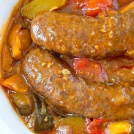 A white bowl contains two pressure cooker sausages submerged in a thick, chunky sauce with visible pieces of red, green, and yellow bell peppers. The sauce has a glossy texture, suggesting a rich, flavorful broth or gravy. The dish is set on a surface with a hint of colorful fabric.