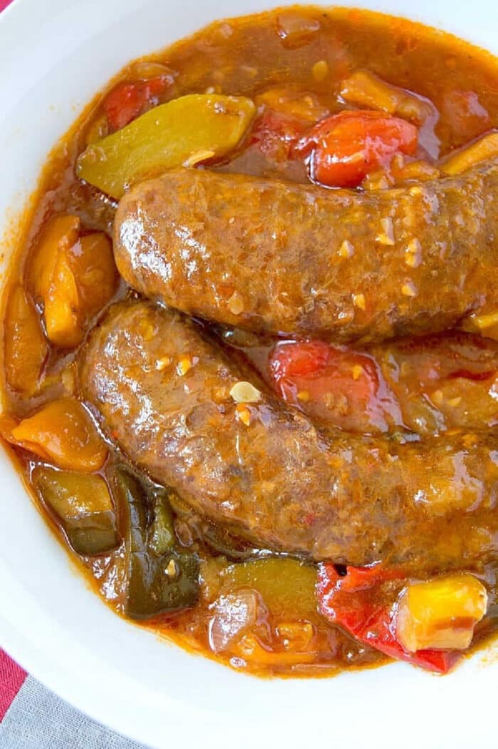 A white bowl contains two pressure cooker sausages submerged in a thick, chunky sauce with visible pieces of red, green, and yellow bell peppers. The sauce has a glossy texture, suggesting a rich, flavorful broth or gravy. The dish is set on a surface with a hint of colorful fabric.