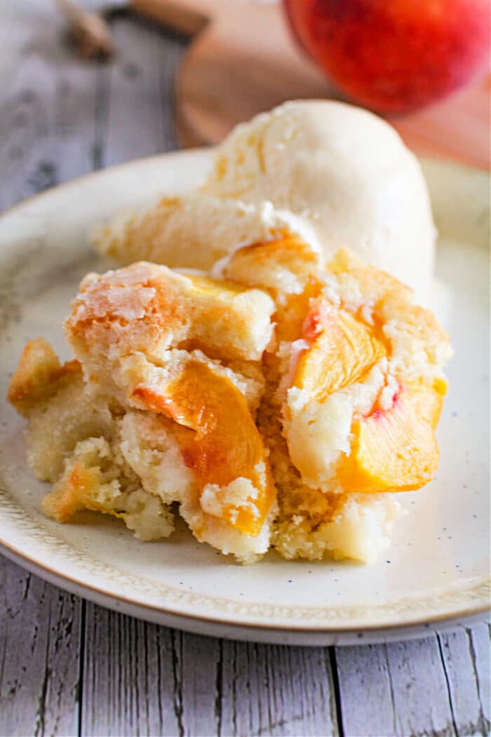 A slice of fresh fruit cake peach cobbler with a scoop of vanilla ice cream graces the plate.