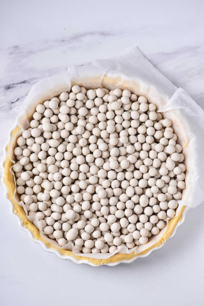 A quiche crust lined with parchment paper, filled with ceramic baking beads on a marble countertop.