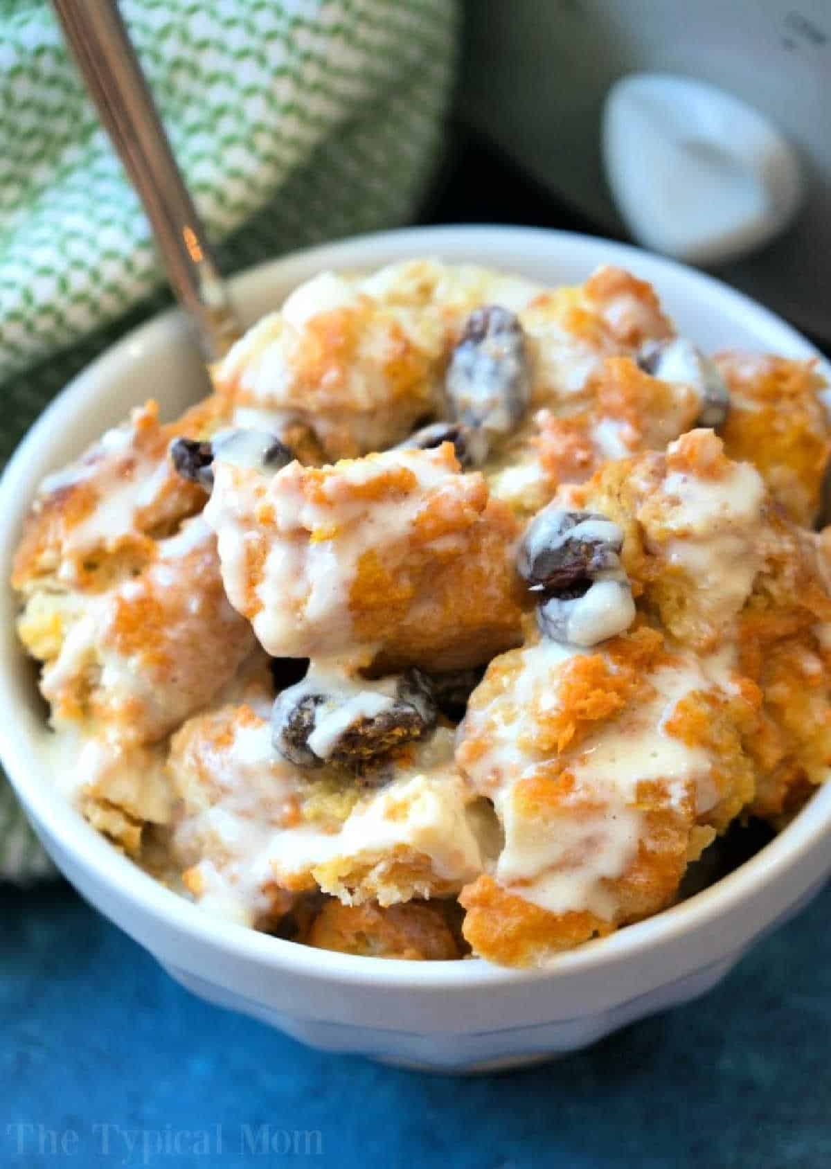 A bowl of sweet crockpot bread pudding with raisins, topped with creamy icing or sauce, sits beside a green patterned cloth.