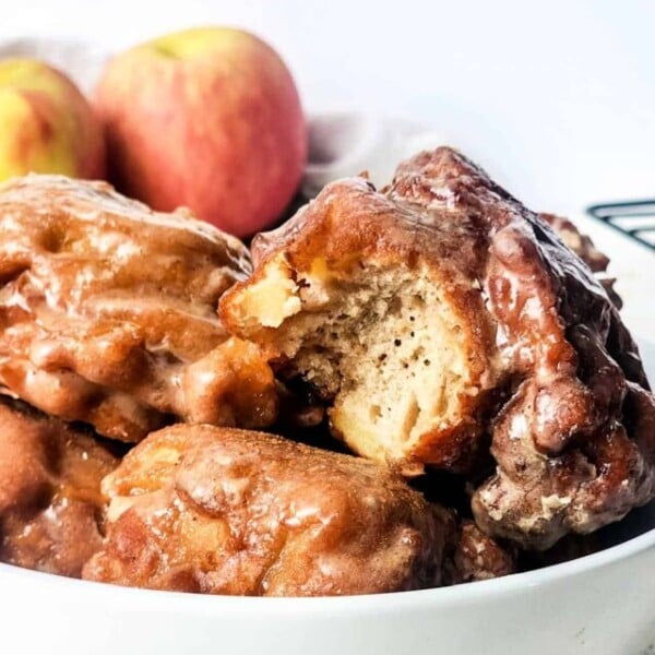 A close-up of a white bowl filled with glazed apple fritters. One fritter has a bite taken out, revealing its moist interior. Two whole apples are blurred in the background. The setting appears to be a light-colored tablecloth.