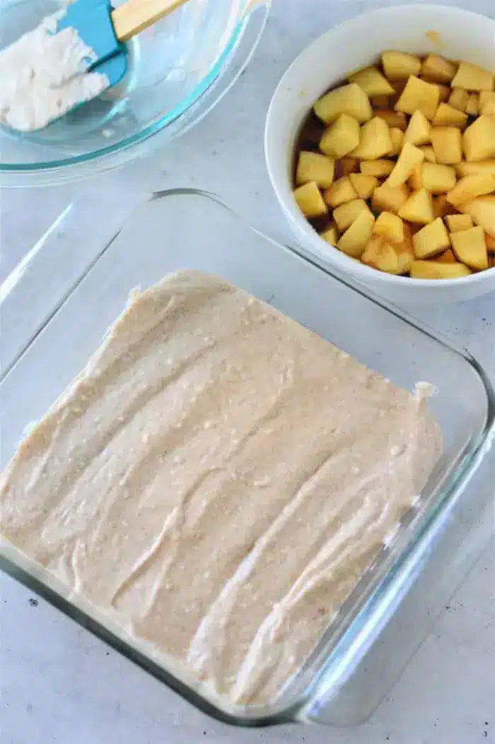 A glass baking dish holds a layer of batter spread evenly. Beside it are two bowls: one with chopped apple pieces and another with a blue spatula resting on the edge, containing a white creamy mixture. This Apple Cobbler with Bisquick is set against a light-colored countertop background.