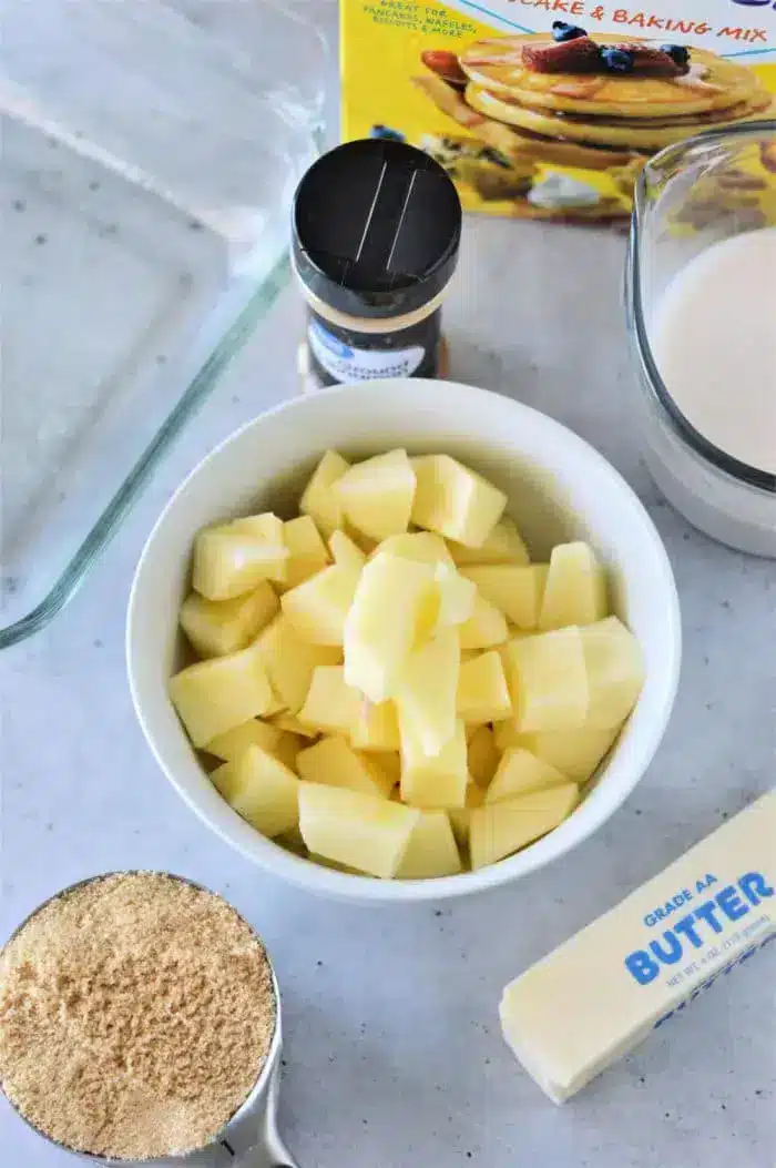 A bowl of diced apples is surrounded by a box of Bisquick for making an apple cobbler, a measuring cup of brown sugar, a stick of butter, a glass of milk, and a bottle of ground cinnamon on a light-colored countertop.