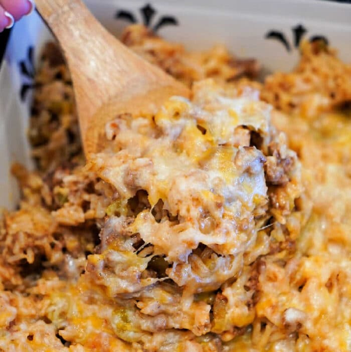 A wooden spoon scoops cheesy, baked hamburger and rice casserole with ground meat from a white dish.