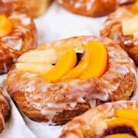 Close-up of delightful puff pastry breakfast creations, glazed and topped with sliced peaches on a pristine white surface.