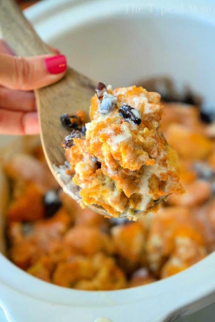 A wooden spoon holds a serving of crockpot bread pudding with raisins above a slow cooker.
