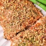         Close-up of almond-crusted salmon fillets garnished with herbs. The salmon is served on a white plate with a few asparagus spears. Overlaid text at the top reads, "Almond Crusted Salmon Recipe.