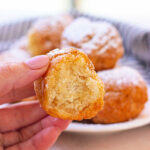 A hand delicately holds a partially eaten fried biscuit donut hole, with a plate piled high with similar treats tempting in the background.