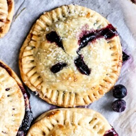 Golden-crusted air fryer blueberry hand pies, topped with sugar crystals and bursting with luscious blueberry filling, rest elegantly on a parchment-lined surface.