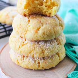 A stack of four delectable cake mix butter cookies, dusted with powdered sugar, sits on a wooden coaster. One has a playful bite taken from it, next to a vibrant green cloth.