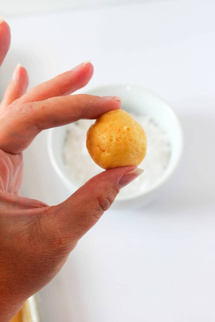 A hand delicately holds a small, round ball of cookie dough over a white bowl on a light background, evoking the satisfying allure of freshly baked cake mix butter cookies.