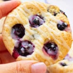 A close-up of a hand holding a round blueberry biscuit with a light icing drizzle. Other blueberry biscuits are visible in the background, along with a few loose blueberries.