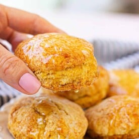 A hand holding a glazed honey biscuit delicately over a plate of assorted biscuits.