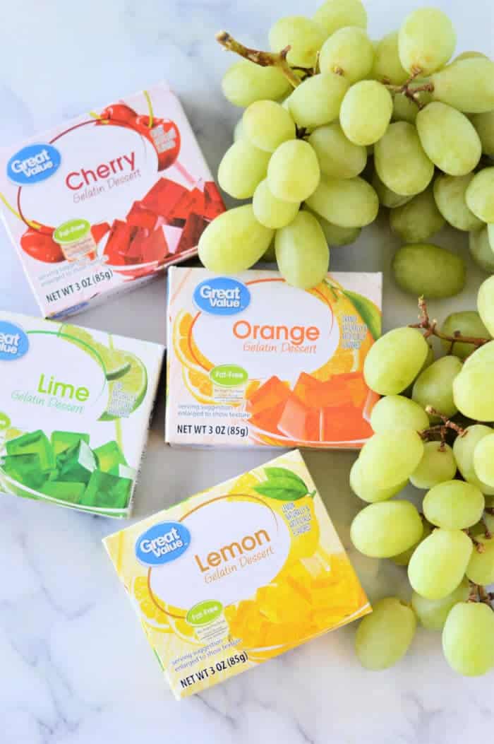 Boxes of gelatin dessert in cherry, lime, orange, and lemon flavors are arranged on a surface along with a bunch of green grapes. Each box is labeled with the flavor and shows colorful gelatin cubes in its respective flavor—perfect for learning how to make candied grapes.