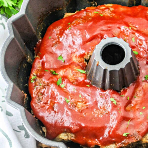 A Bundt Pan Meatloaf glistens under a glossy tomato sauce, with fresh parsley leaves artfully sprinkled on top. In the background, a bunch of parsley rests beside a patterned cloth, complementing this culinary centerpiece.