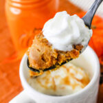 A spoonful of pumpkin mug cake with whipped cream sits invitingly atop a white mug, accompanied by an orange jar.