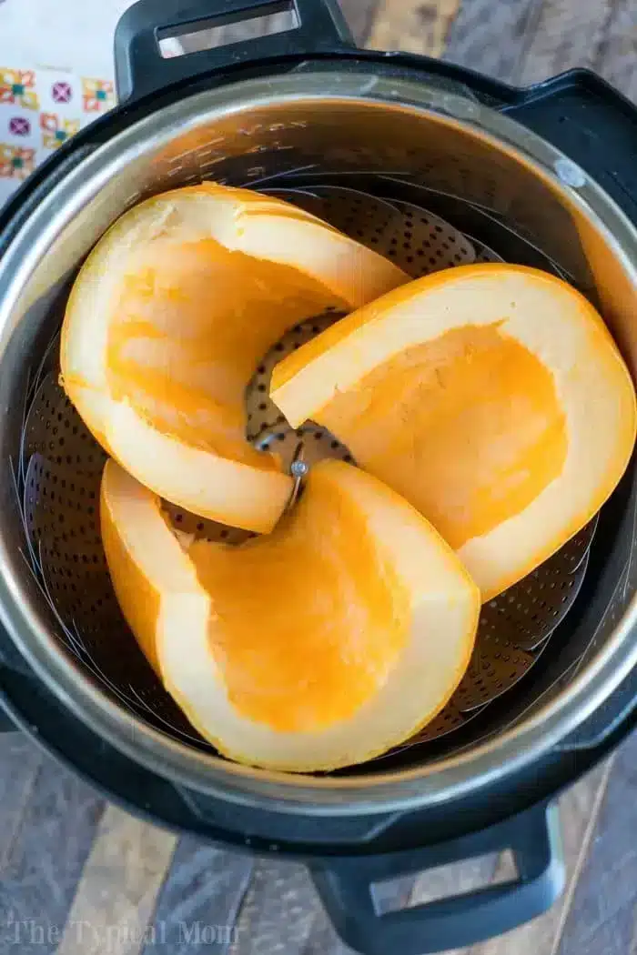 Three partially peeled and deseeded pumpkin quarters are placed in a steamer basket inside an Instant Pot. The setup appears ready for cooking, with the promise of air fryer pumpkin seeds from the removed seeds.