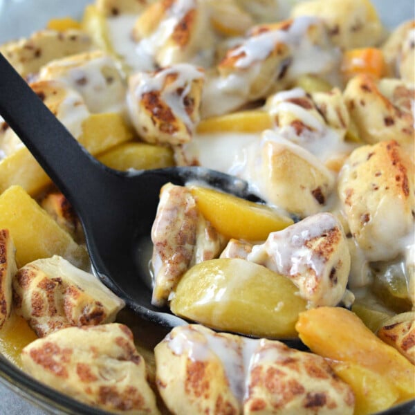 A skillet of cinnamon rolls with peaches and cream, served with a black spoon, offers a delightful twist reminiscent of a cinnamon roll peach cobbler.