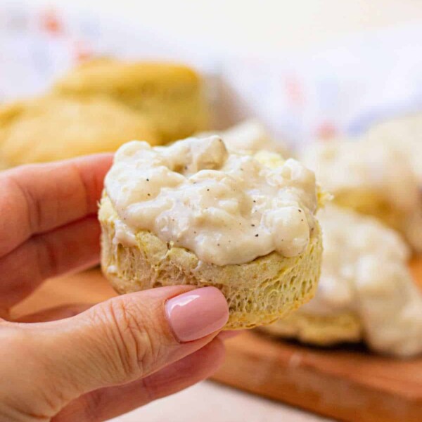 A hand holds a biscuit topped with creamy white vegan gravy, with more vegan biscuits in the background.