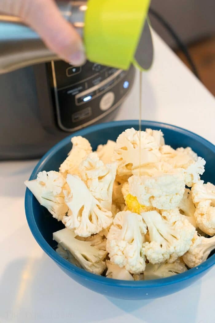 Easy air fryer cauliflower is easy to make in your Ninja Foodi or other machine. Healthy and full of flavor it's a great vegetable side dish or snack. #airfryer #ninjafoodi #cauliflower #vegetables #vegetarian #sidedish Olive oil is being poured onto fresh cauliflower florets in a blue bowl, perfect for making crispy air fryer cauliflower near an electric cooker.