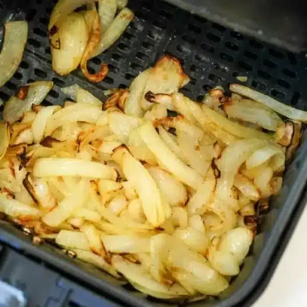 Sliced onions are shown cooking in an air fryer basket, partially browned and in the process of caramelizing. The air fryer is open to display the air fryer onions inside.