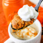 A close-up image of a black spoon holding a bite of Pumpkin Mug Cake topped with whipped cream. In the background, an orange ceramic container and a white mug brimming with more Pumpkin Mug Cake and whipped cream sit on an orange surface, creating a cozy autumnal scene.