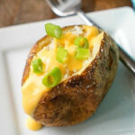 Crockpot baked potatoes topped with cheese sauce and chopped green onions on a white plate with a fork.