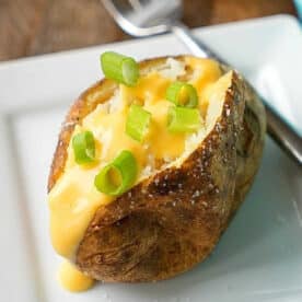 Crockpot baked potatoes topped with cheese sauce and chopped green onions on a white plate with a fork.