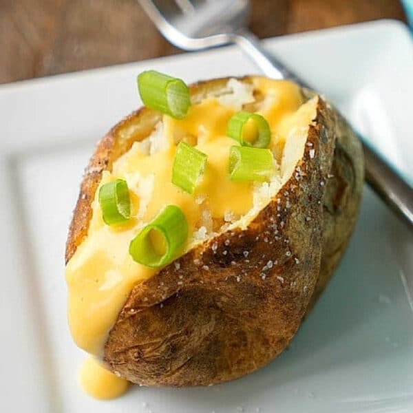 Crockpot baked potatoes topped with cheese sauce and chopped green onions on a white plate with a fork.