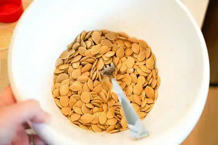 A white bowl filled with a generous amount of air fryer pumpkin seeds is shown. A fork is resting in the bowl, partially covered by the seeds. The image focuses on the tan-colored seeds, which are evenly spread out in the bowl.