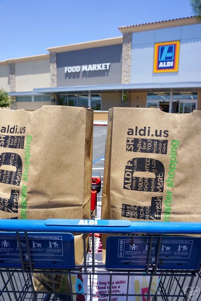 A shopping cart filled with brown paper bags is in the foreground, with the logo "aldi.us" visible on the bags. An Aldi store exterior is visible in the background, featuring a "Food Market" sign accompanying the Aldi logo. Don't forget to grab your free printable meal planner for an organized shopping experience!