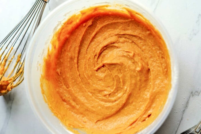 A bowl of creamy pumpkin cake mix with a whisk sits on a white marble countertop, ready to transform into delightful mini bundt cakes.