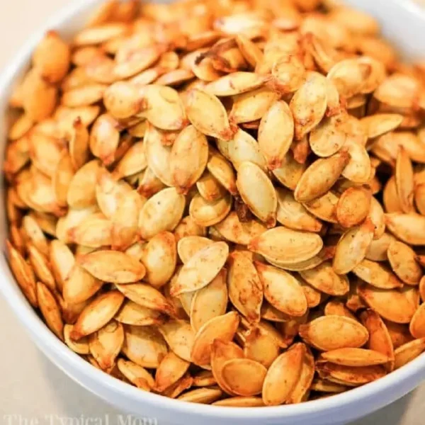 A white bowl filled with a large quantity of seasoned, roasted air fryer pumpkin seeds. The seeds appear golden brown and are seasoned with various spices.