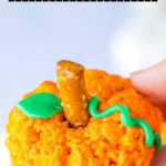 A hand holds a festive Halloween Rice Krispie Treat: an orange, pumpkin-shaped delight complete with a pretzel stem and green icing leaves.