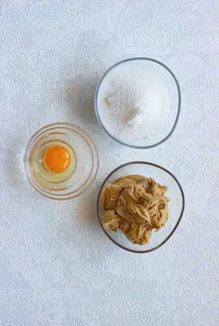 Three clear bowls on a textured white surface: the top bowl contains granulated white sugar, the bottom left bowl holds a raw egg, and the bottom right bowl brims with creamy peanut butter—perfect for making Peanut Butter and Grape Jelly Cookies.