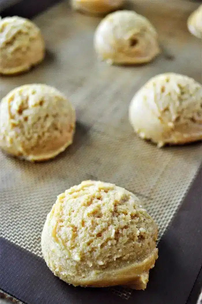 Scoops of unbaked Peanut Butter and Grape Jelly cookie dough are placed on a baking sheet covered with a silicone mat. The dough appears slightly textured and is evenly spaced apart, with some of the scoops in the foreground and others in the background.