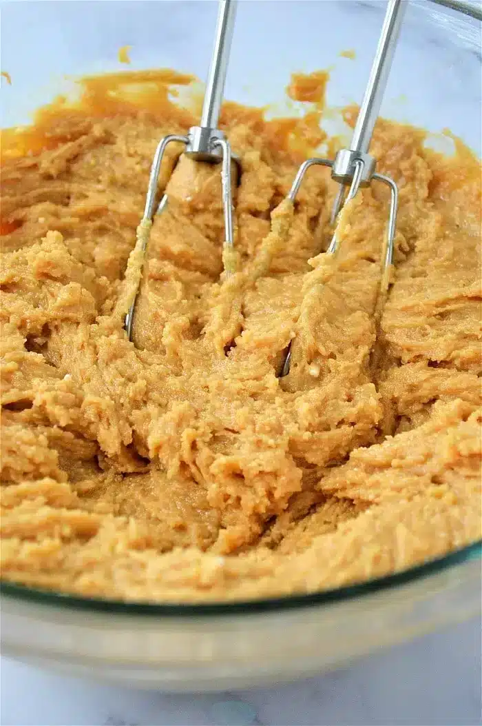 A close-up view of a glass bowl filled with cookie dough for Peanut Butter and Grape Jelly Cookies being mixed by a hand mixer with two beaters. The light brown dough appears smooth and creamy.