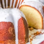 A sliced 7-up pound cake bundt cake is sitting on a white plate. The moist yellow interior and golden brown crust are beautifully complemented by a thin white glaze. The plate rests on a floral tablecloth, adding to the charm of this classic dessert.