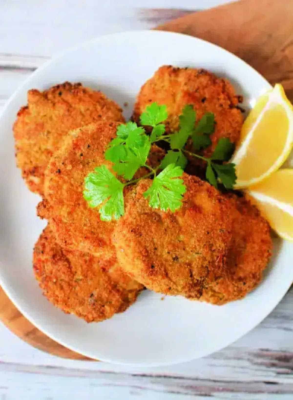 Five golden brown Air Fryer Crab Cakes garnished with cilantro and lemon wedges on a white plate.
