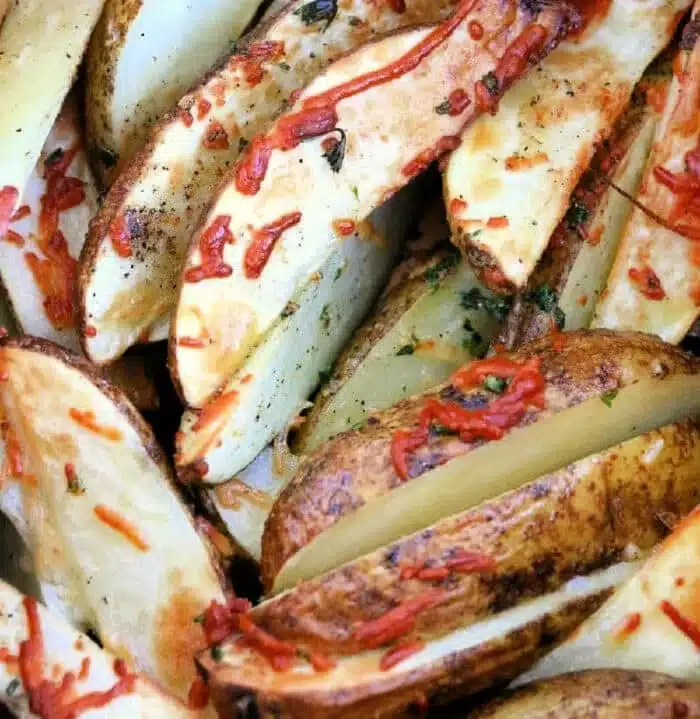 Close-up of air fryer potato wedges topped with shredded cheese and herbs. The wedges are golden brown and appear crispy, with visible seasoning flecks and melted cheese.