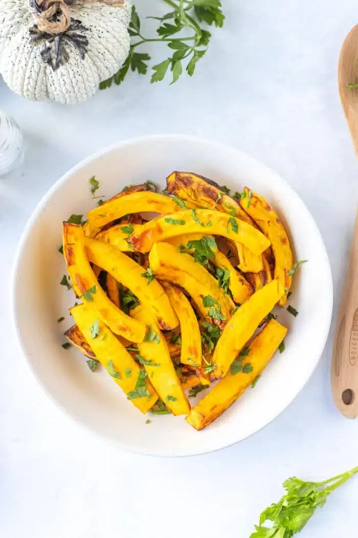 A white bowl contains air fryer roasted delicata squash slices garnished with chopped parsley. A wooden spoon and a small white pumpkin are partially visible in the background. The bowl sits on a white surface.