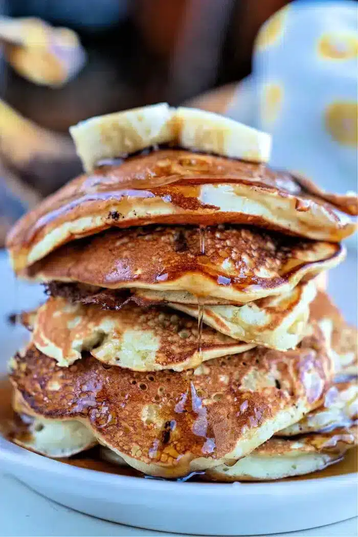 A close-up image of a stack of banana chocolate chip pancakes on a white plate. The pancakes are drizzled with syrup and topped with a slice of butter. The background is blurred, focusing attention on the delectable pancakes.