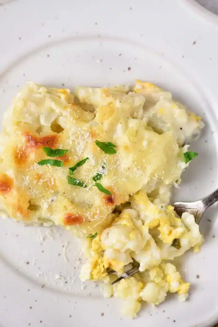 A piece of Cheesy Cauliflower Casserole is served on a white plate. The casserole is topped with melted cheese and garnished with chopped parsley. A metal fork is placed next to the casserole, picking up a small portion of it.